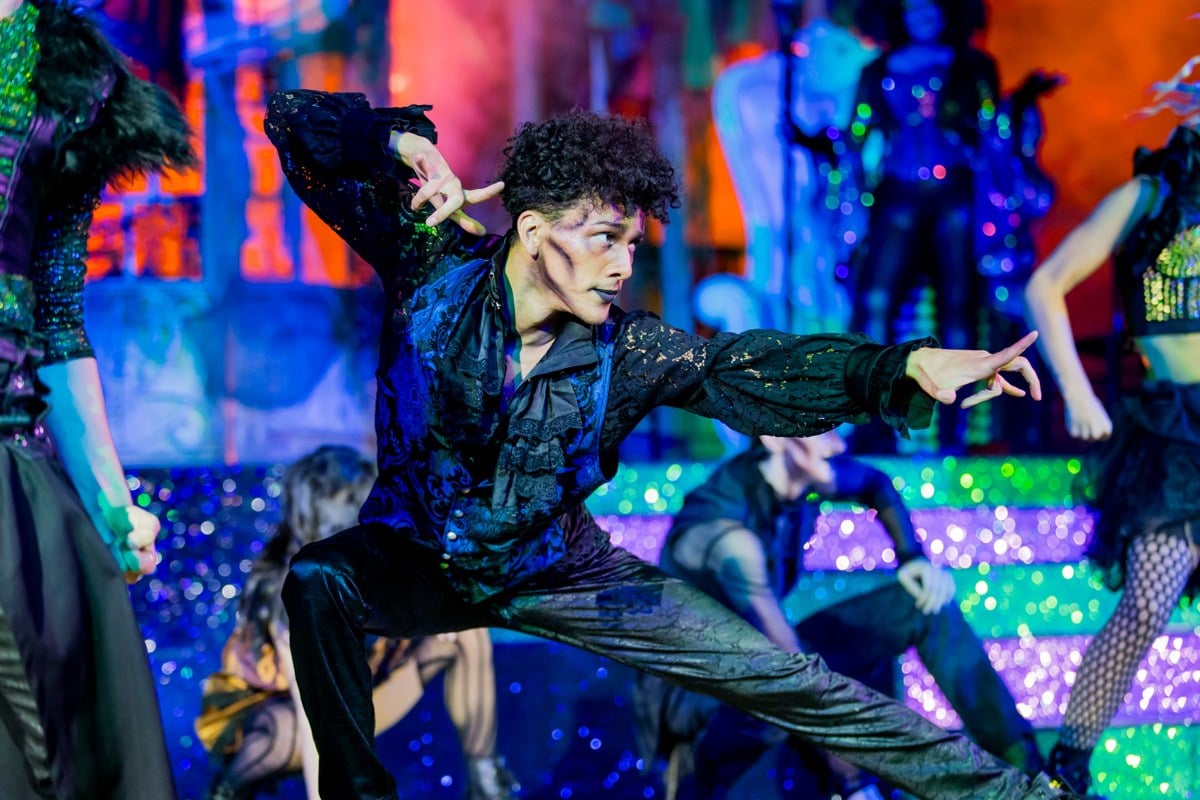 A dancer in a sequined jacket leaps across a brightly lit stage with other performers behind him