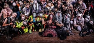 Dozens of costumed actors in monster makeup pose together on a wooden stage at night, crouching and reaching toward the camera