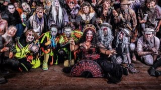 Dozens of costumed actors in monster makeup pose together on a wooden stage at night, crouching and reaching toward the camera