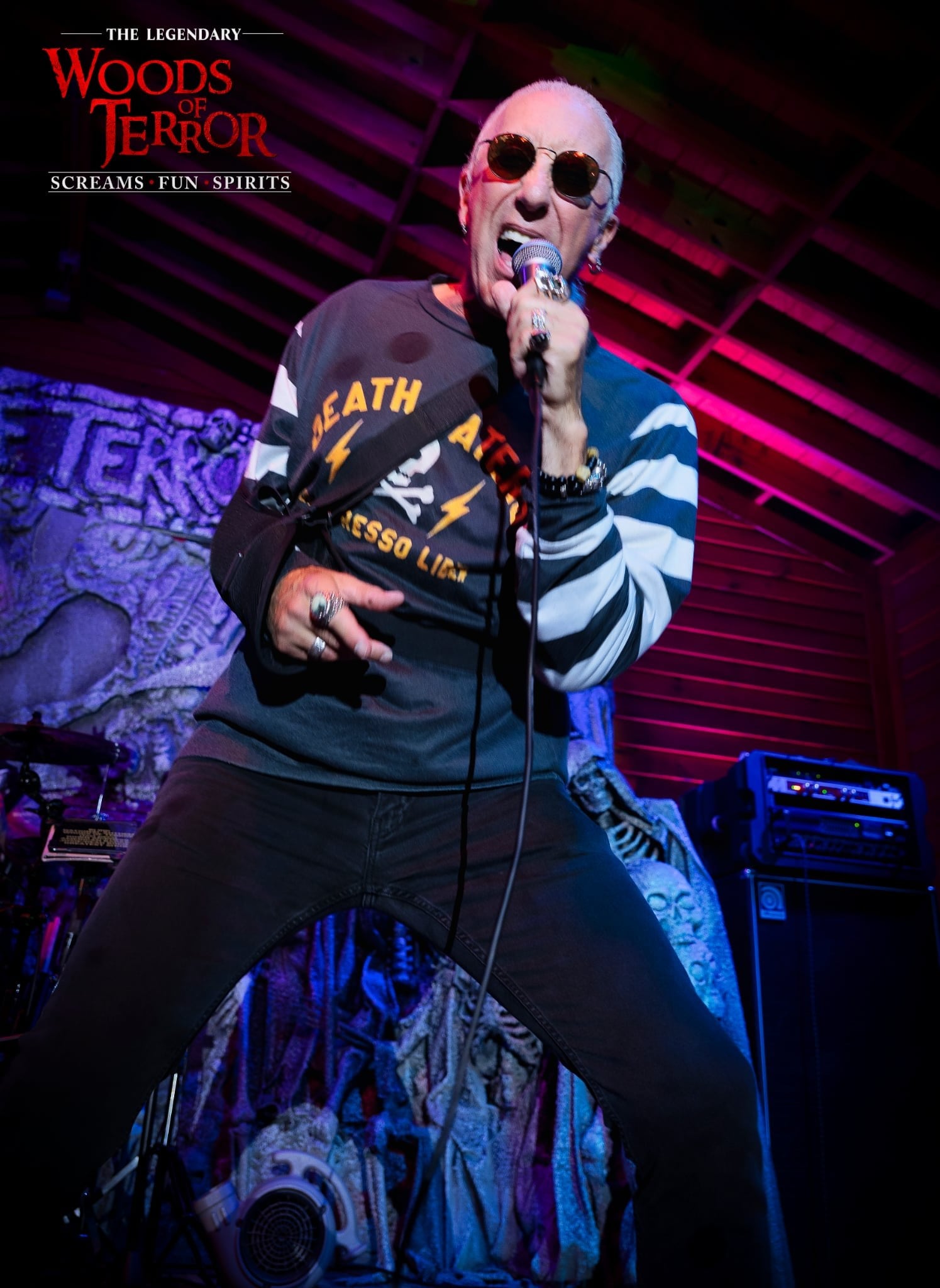 Dee Snider sings into a microphone on a stage, wearing sunglasses and a striped shirt, under red and purple light