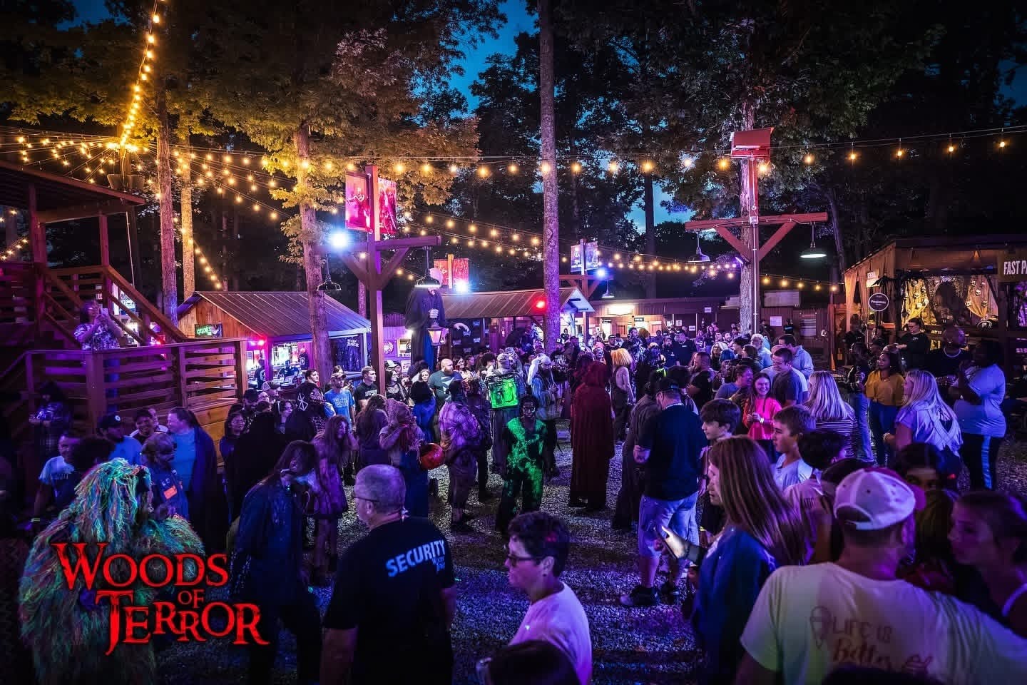 A crowd fills a gravel midway under strings of lights, with wooden buildings and trees around it at dusk