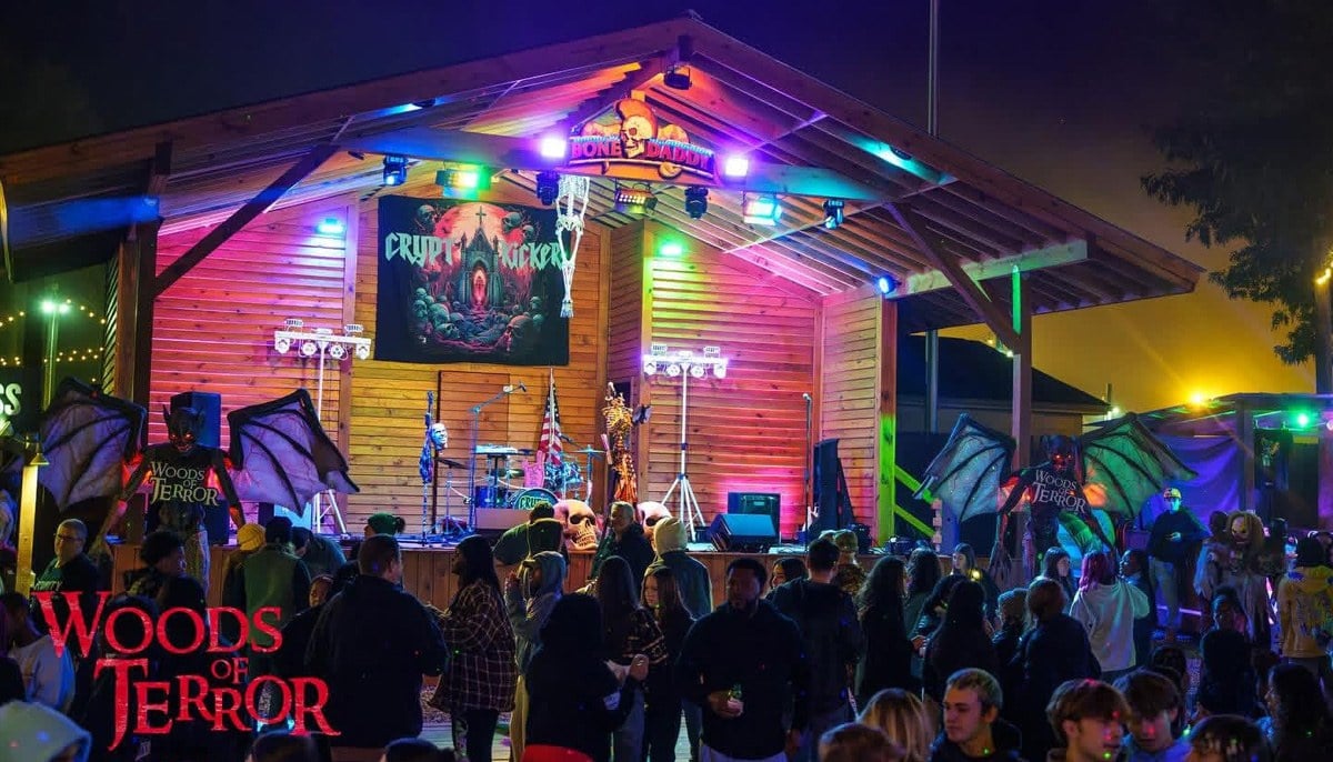 A crowd stands in front of a wooden stage lit in purple and green, with a band playing and winged monster statues on either side